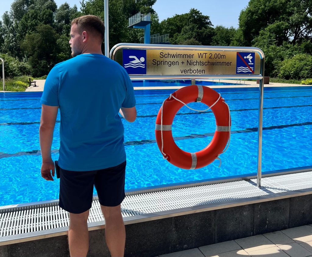 Ein Schwimmmeister steht mit dem Rücken zur Kamera vor einem Schwimmbecken . Neben ihm hängt ein Rettungsring
